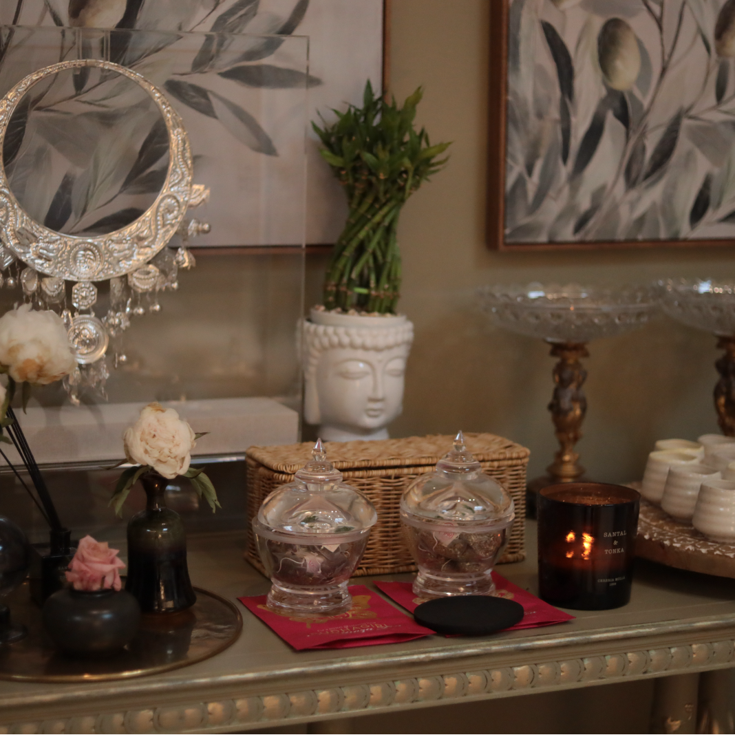 A styled interior tabletop scene with two clear glass lidded jars as the apparent products, placed on red cards beside a black candle, flowers, a wicker basket, and decorative wall art.