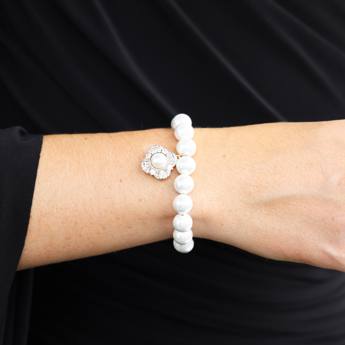 BuDhaGirl white pearl bangle bracelet for women with a delicate floral silver charm worn on a wrist against black fabric