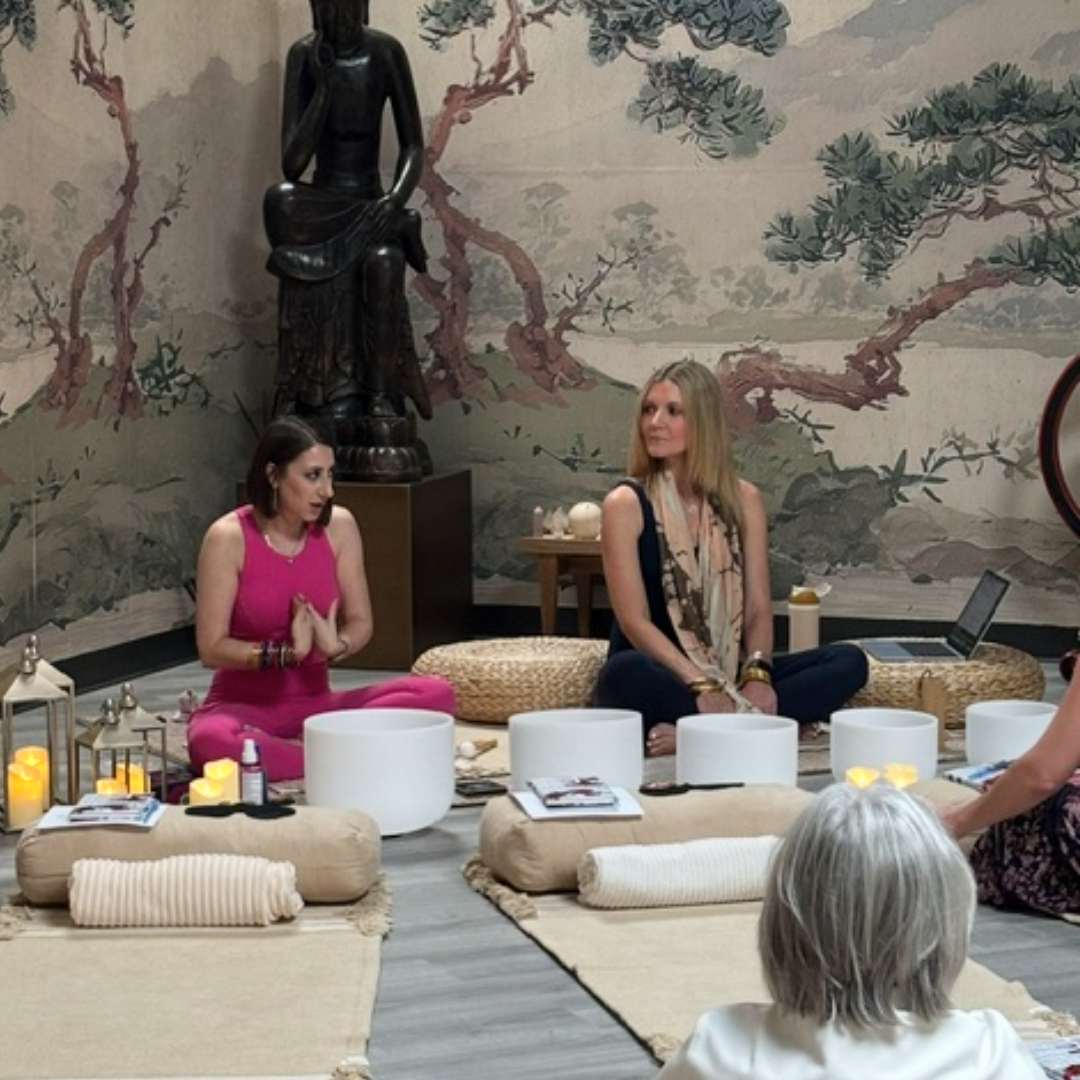 Two women meditate in a serene room with a large Buddha statue, traditional wall mural, and white sound bowls. One woman wears a bright pink outfit, and the other wears dark clothing with a patterned scarf.