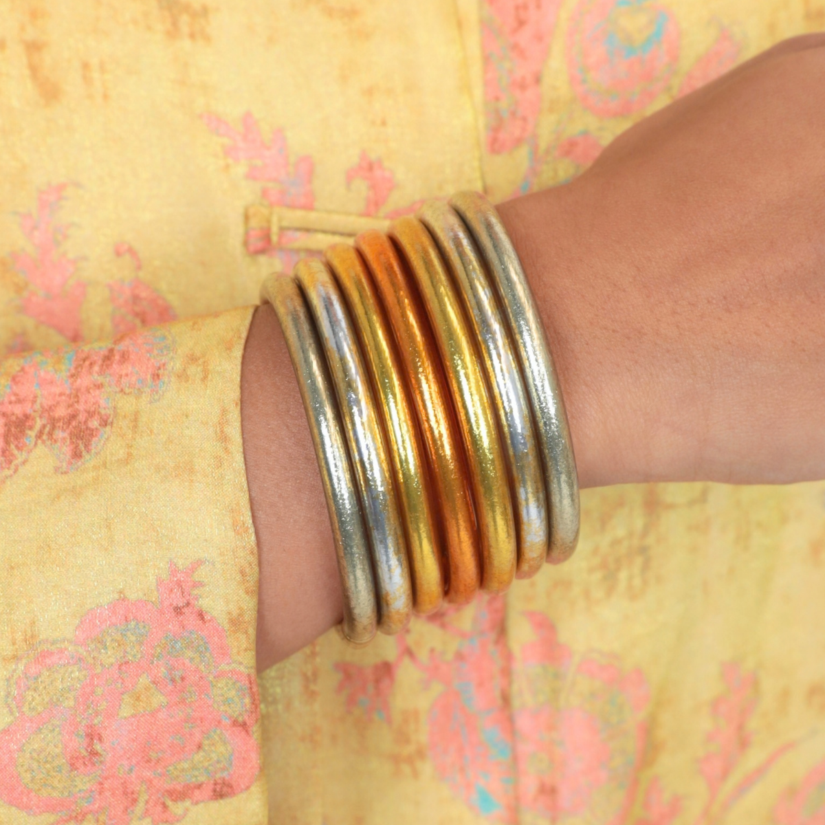 BuDhaGirl gold and silver bangle bracelets for women stacked on a wrist against a soft yellow floral garment