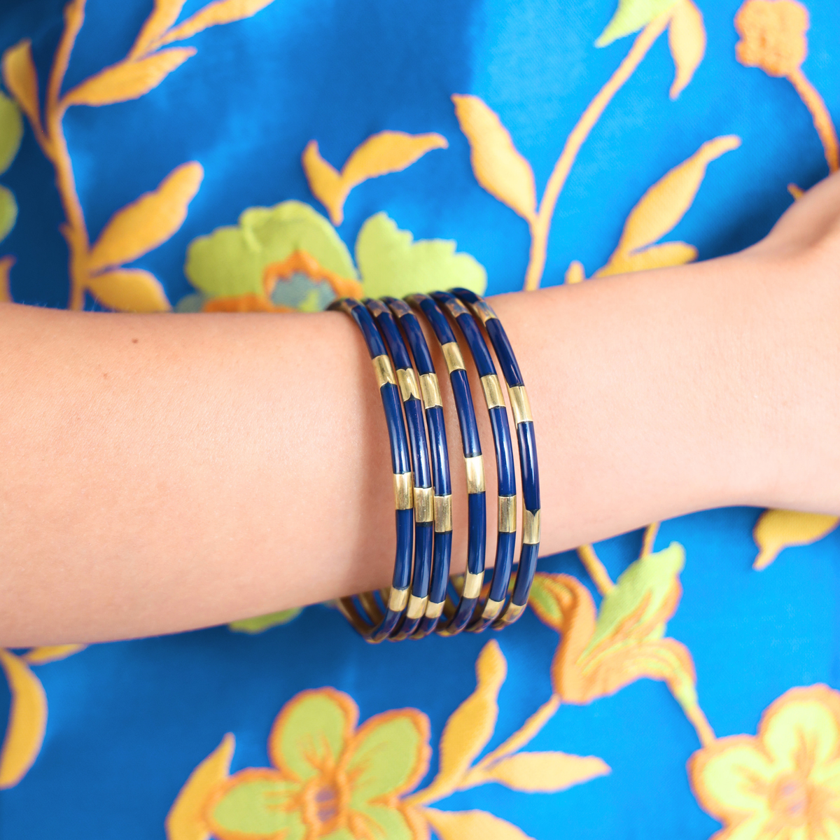 A stack of multiple bangle bracelets with dark blue and gold striped patterns, worn on a person's wrist against a bright blue floral fabric background.