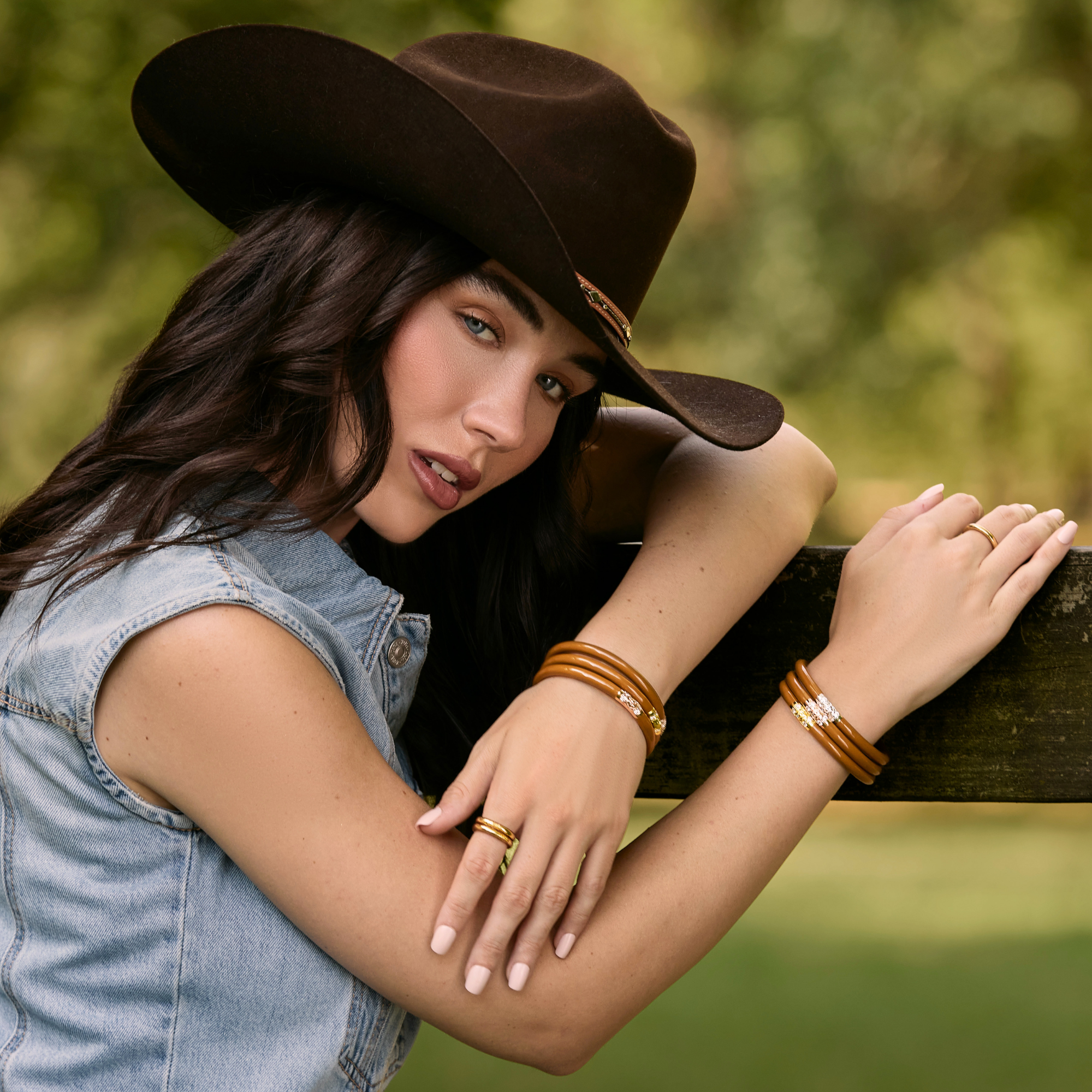 A woman wearing a brown wide-brim hat and a light blue denim vest leans on a wooden fence outdoors, wearing multiple gold-toned bracelets and rings.