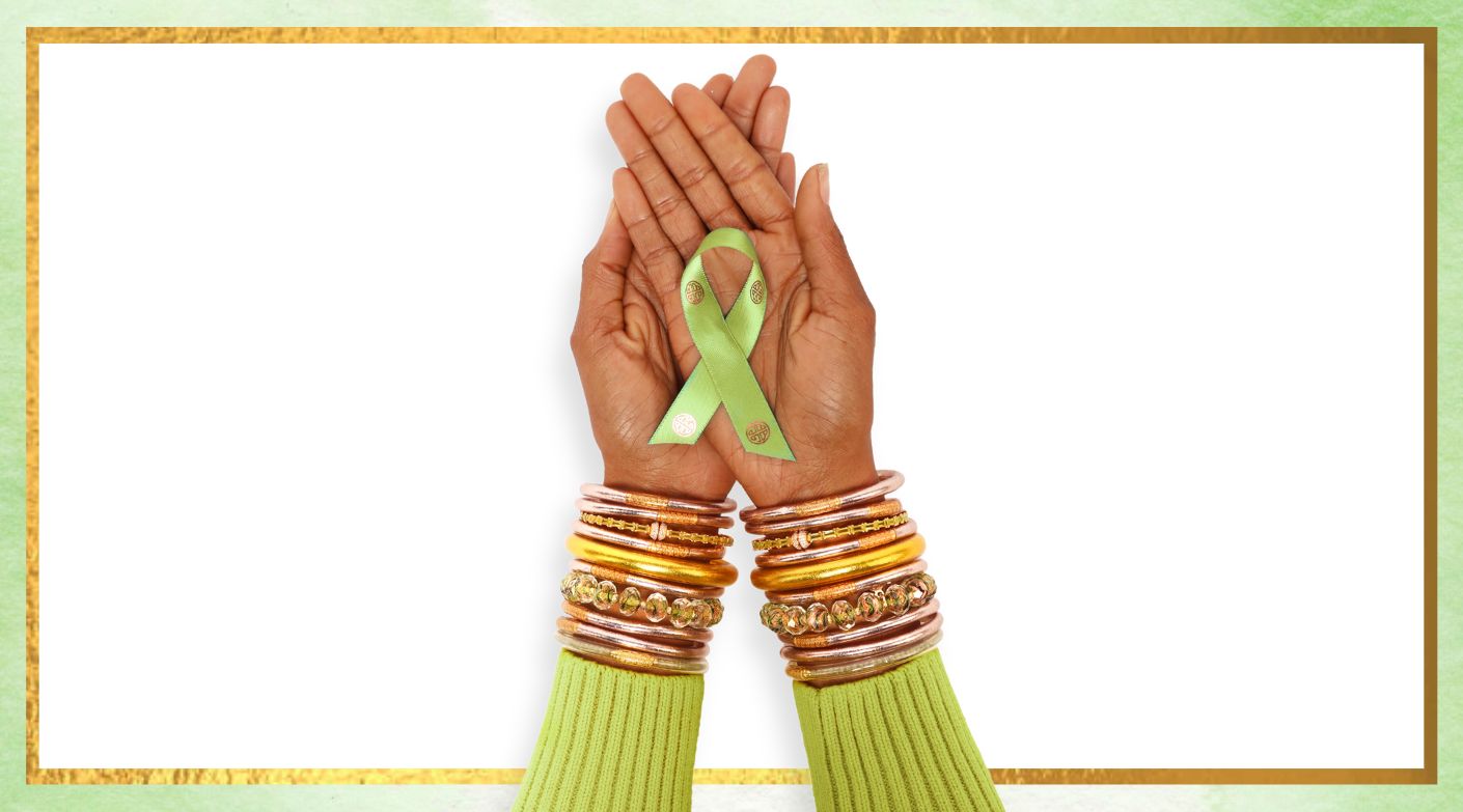Woman in green sweater with hands crossed with green mental health awareness ribbon in hand, wearing BuDhaGirl Rose Gold AWB, Aurora Peridot crystal bracelet, Gold Tzubbie bangle bracelet, Pond Bracelet and Peridot Luxe AWB | BuDhaBrief by BuDhaGirl