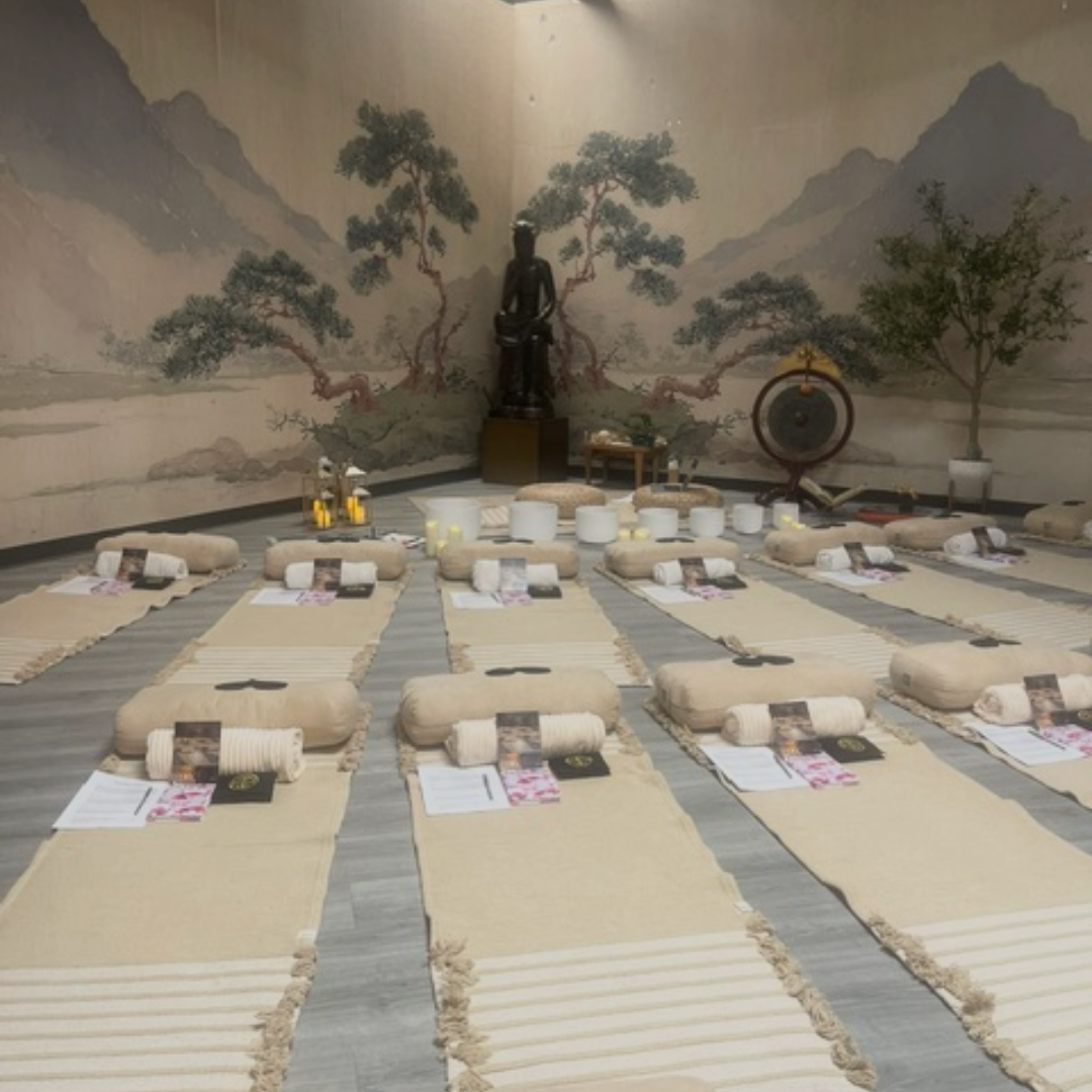 A serene meditation or spa room with multiple beige mats arranged in rows on a gray floor, each styled with a rolled towel, booklet, and small tray. The room has a large wall mural depicting mountains and trees, a black statue, and a decorative drum.