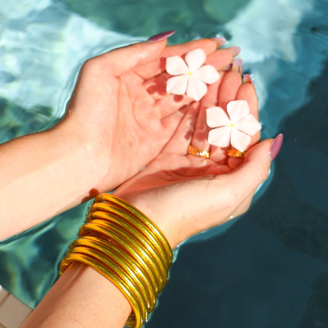 Woman with Hands in Water Holding Flowers and Wearing Gold All Weather Bangle Bracelets | BuDhaGirl