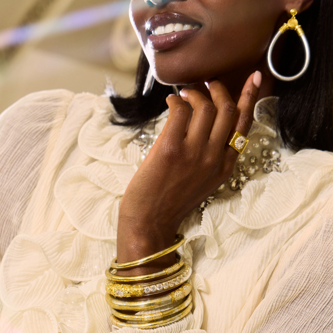 Close up of a woman wearing layered gold bangle bracelets and a square ring with large teardrop earrings on a cream ruffled blouse