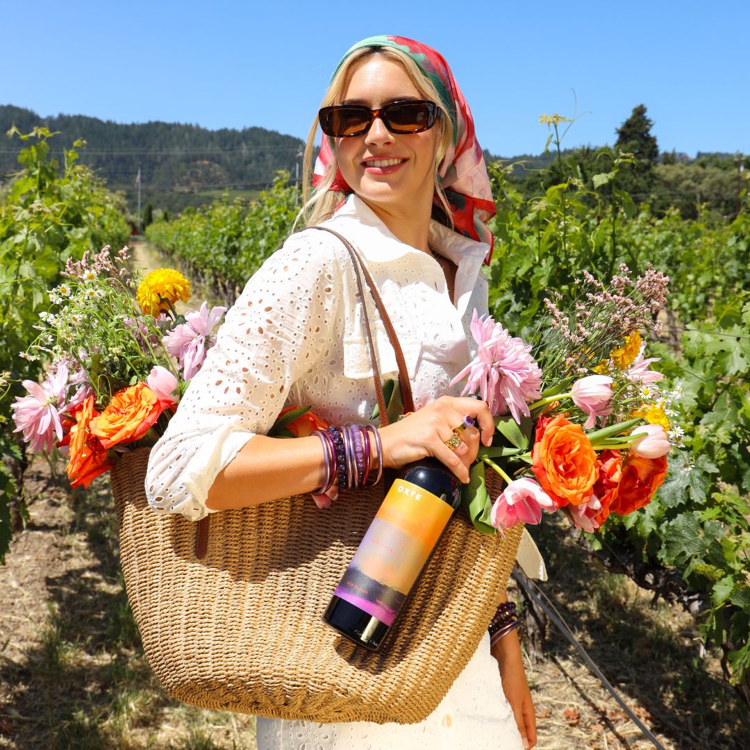 Woman in Vineyard, Holding a Raffia Tote Bag Filled with Flowers and a Bottle of Wine While Wearing a Bangle Bracelet Stack | BuDhaGirl