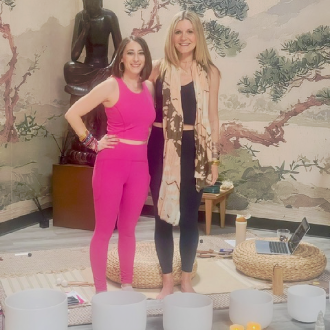 Two women standing in a decorated room with a large Buddha statue and landscape wall mural. The woman on the left wears a bright pink activewear set, and the woman on the right wears a black top and leggings with a beige and brown patterned scarf.