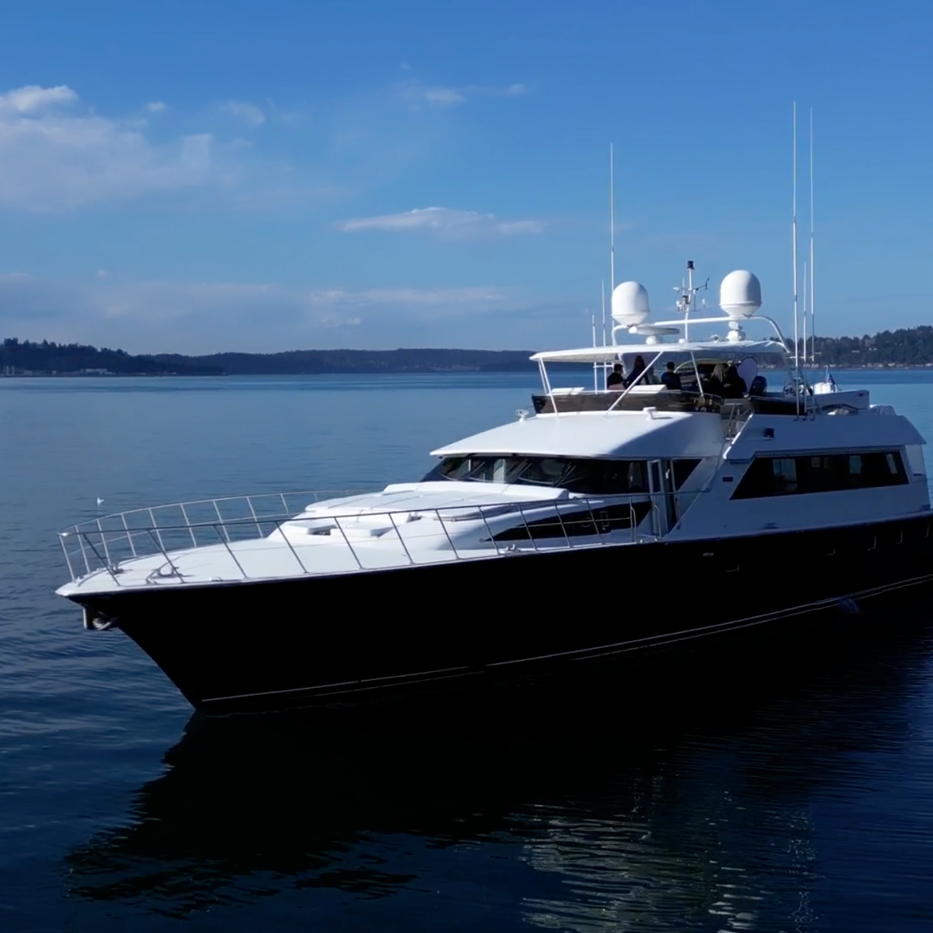 A luxury motor yacht with a white upper deck and black hull, floating on calm blue water under a bright blue sky with scattered clouds.