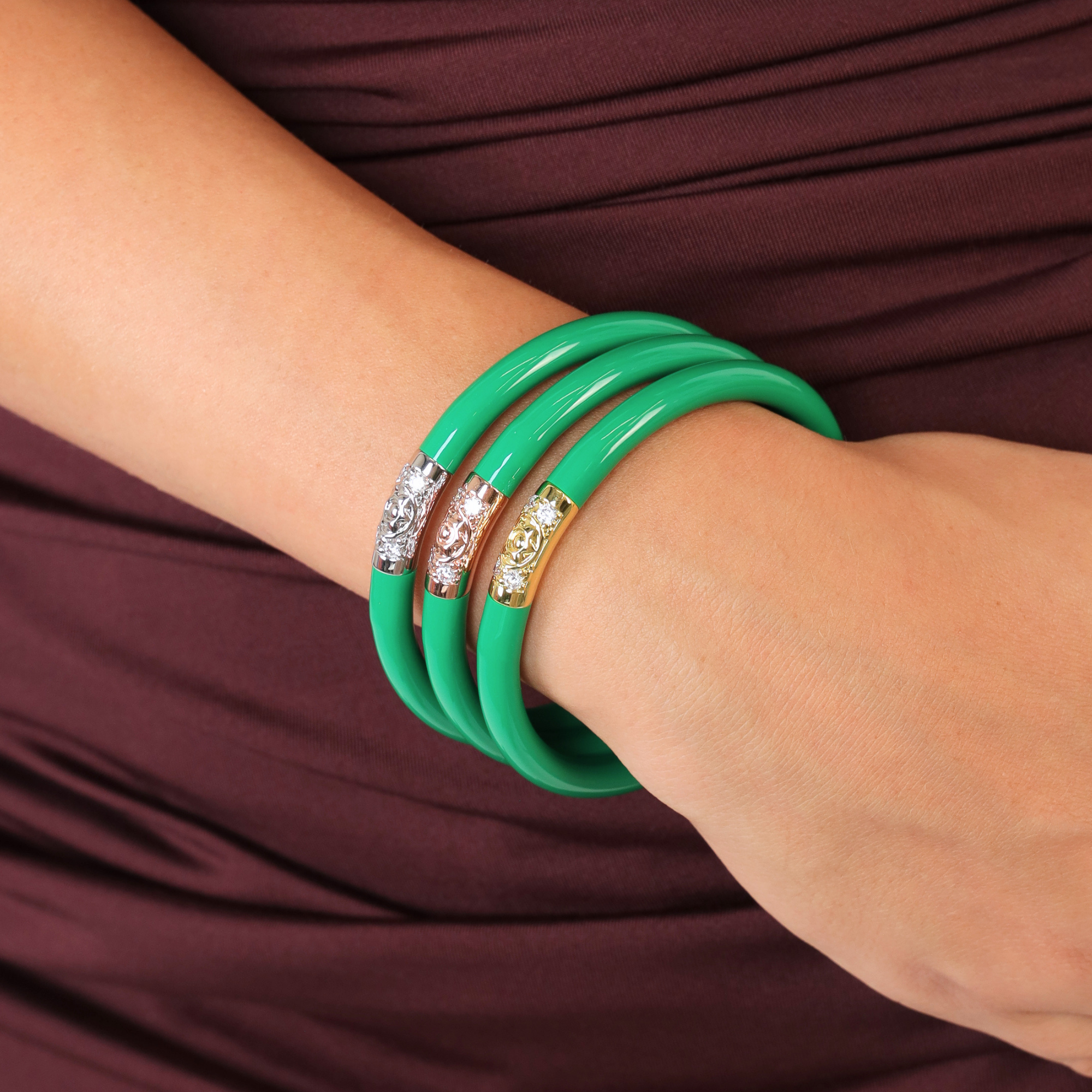 Three glossy green bangle bracelets with decorative crystal-encrusted bands in silver, rose gold, and yellow gold, shown worn on a person's wrist against a dark burgundy dress.