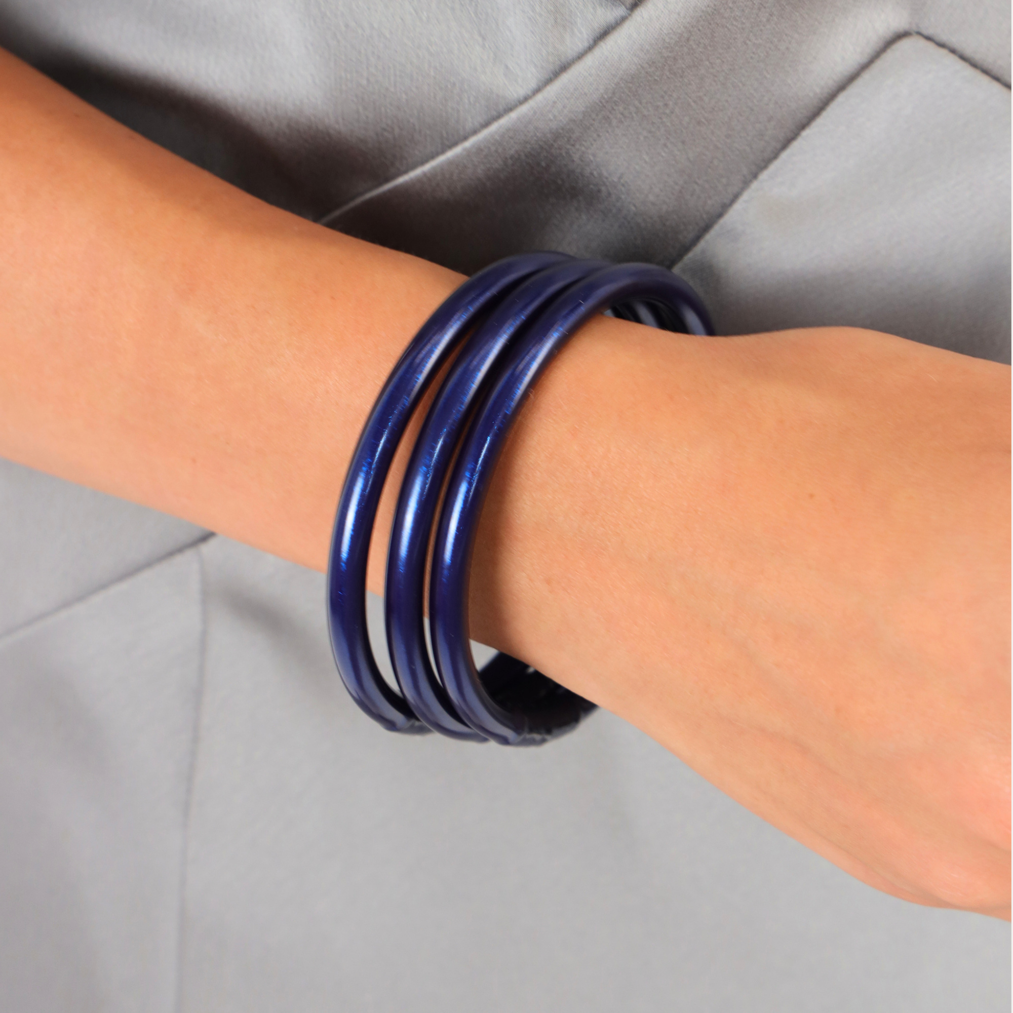 A close-up of a person's wrist wearing four glossy dark blue bangles stacked together, with a light gray blazer in the background.