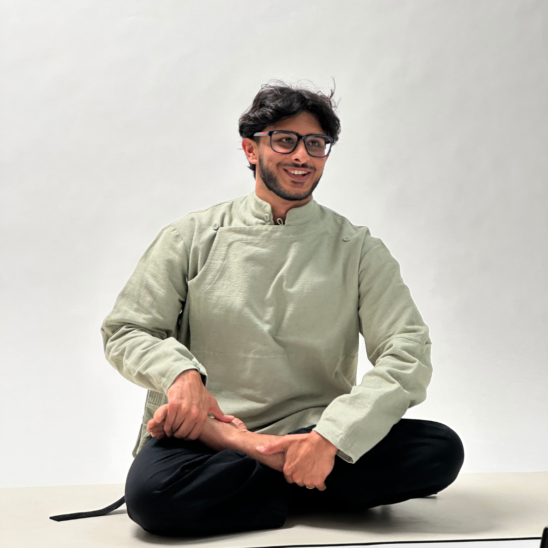 A man wearing a light sage green traditional long-sleeve shirt and black pants sits cross-legged on a white studio floor against a light gray background.