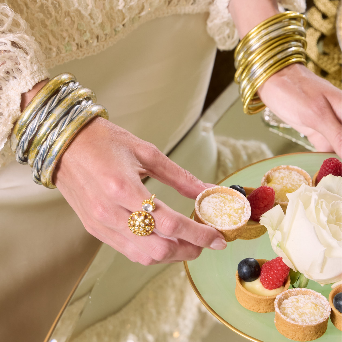 Close-up of a woman wearing stacked gold and silver bangle bracelets while holding a powdered mini tart beside a plate of pastries and berries