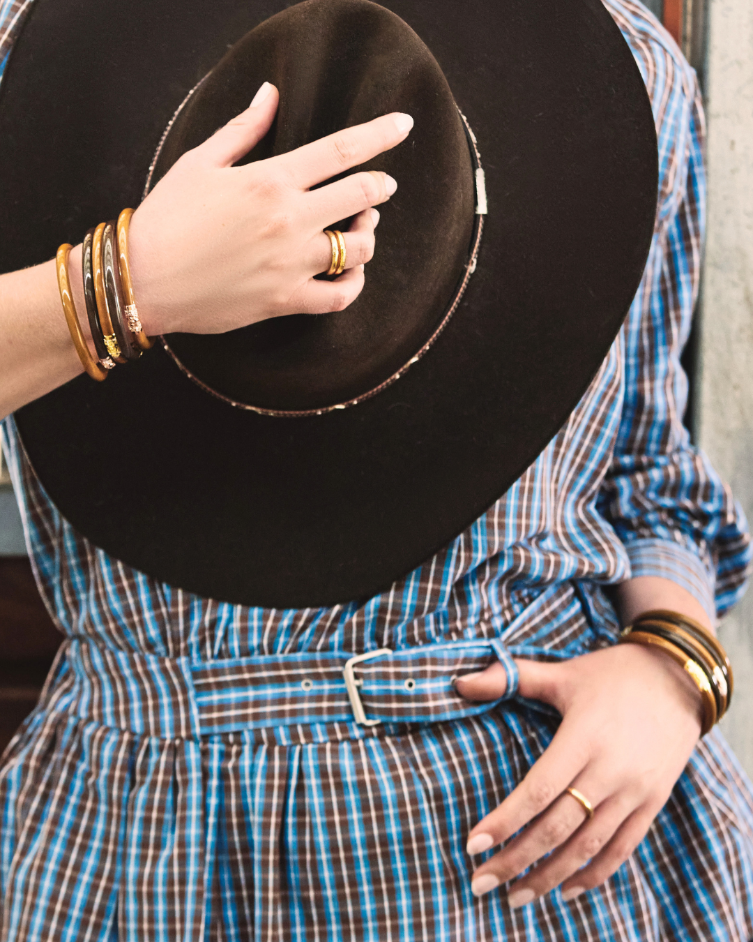 A close-up lifestyle shot of a person wearing a blue and brown plaid shirt dress with a matching belt, holding a black wide-brim hat. The person is accessorized with multiple gold bangles and rings.