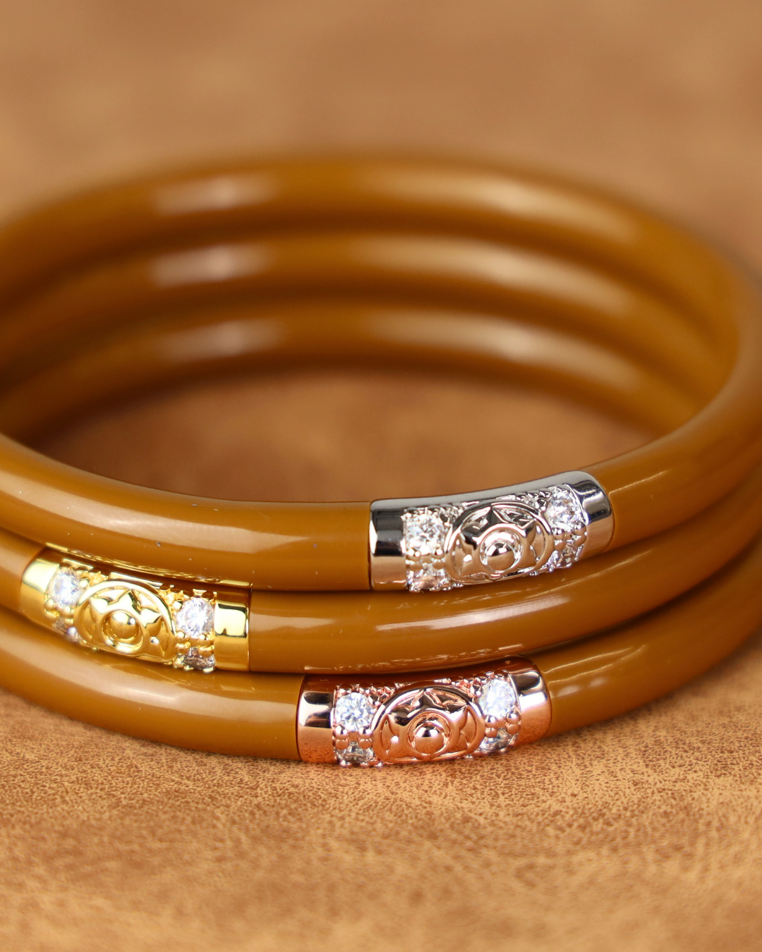 Three stacked brown bangle bracelets with decorative silver-toned and rose gold end caps featuring clear rhinestones, set against a textured brown background.