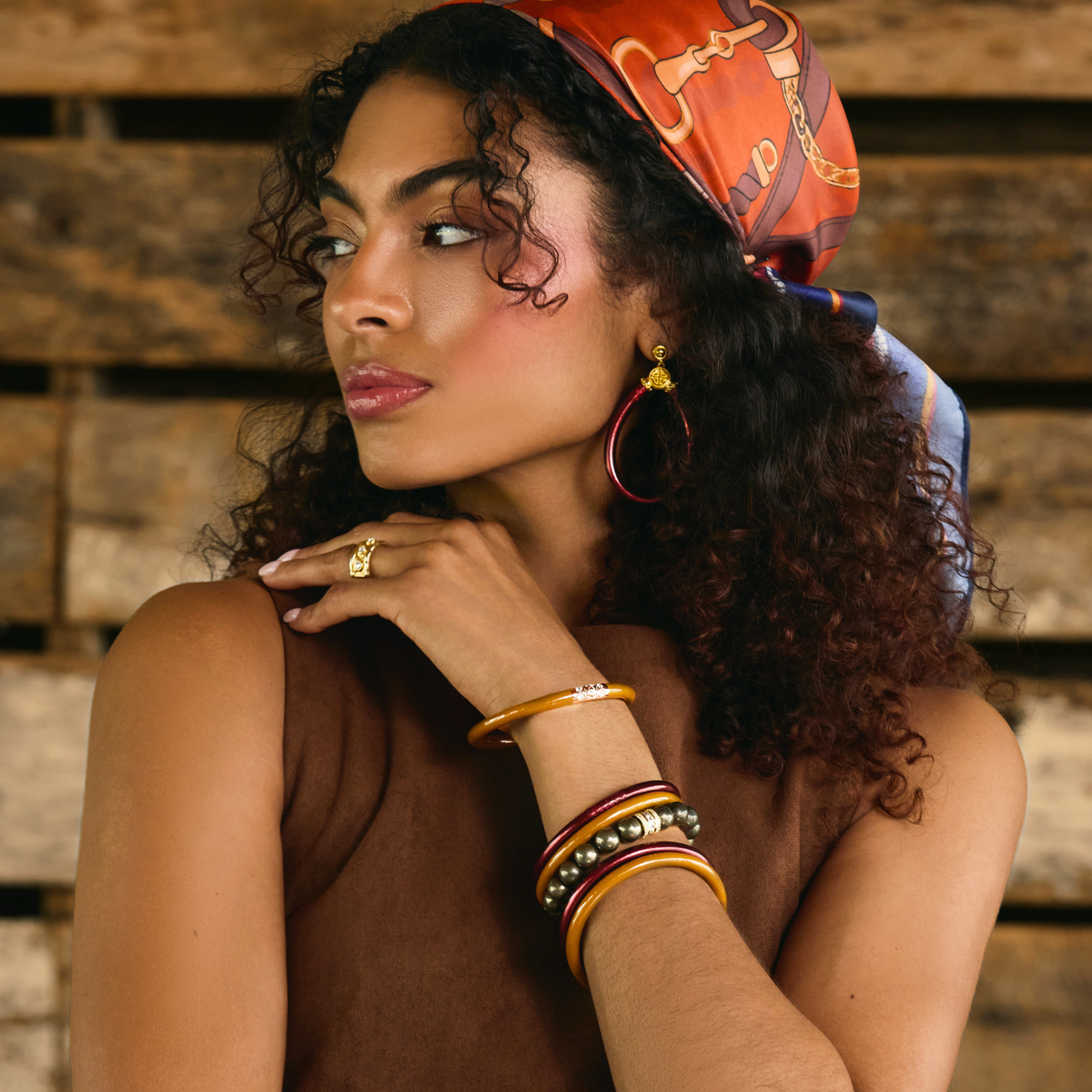 A woman with curly dark hair wears a patterned orange and red headscarf, teardrop merlot all weather earrings, a gold ring, and stacked gold and red bracelets, posed against a rustic wooden plank background.