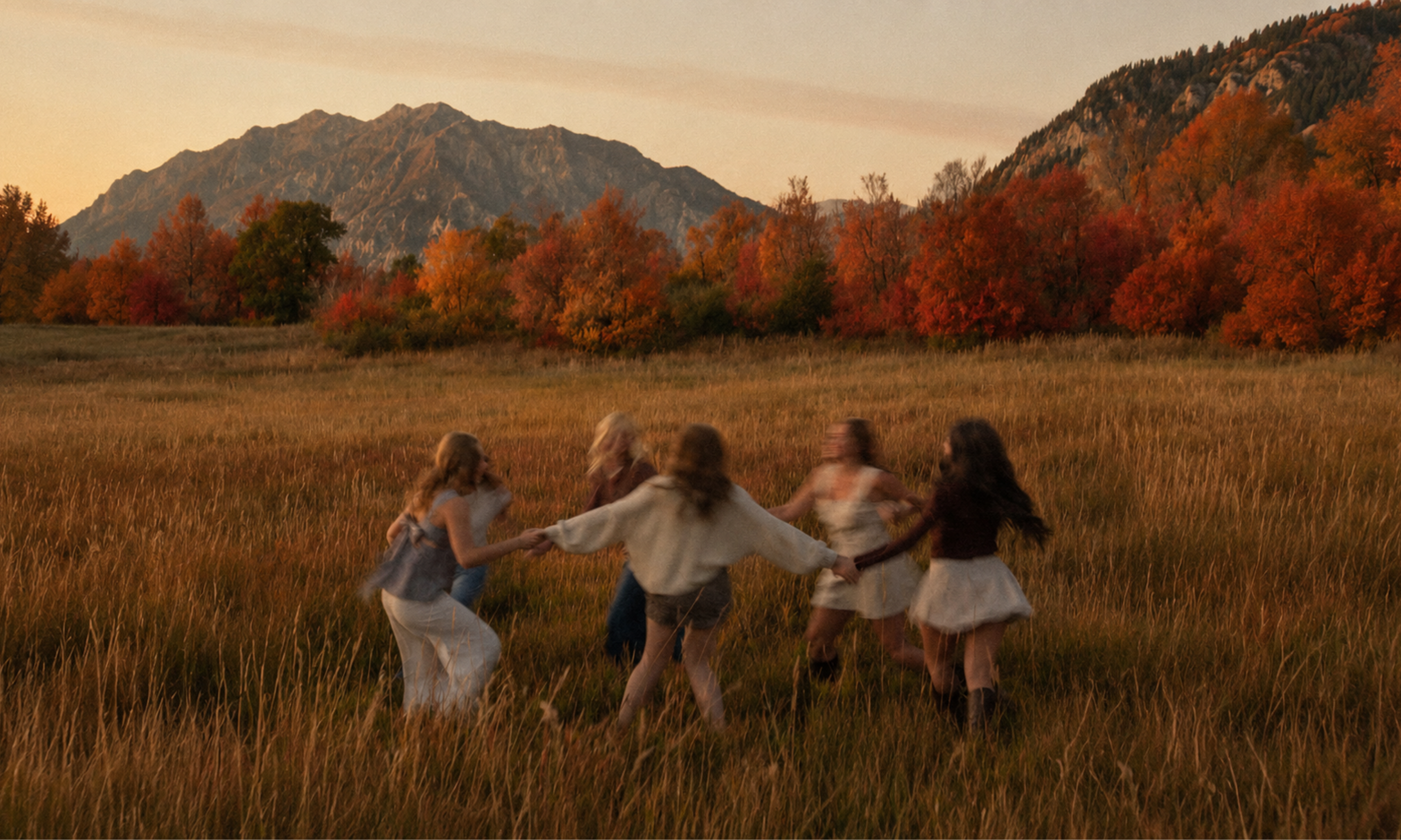 Group of girls joyfully dancing in a scenic autumn field with colorful trees and mountains in the background.